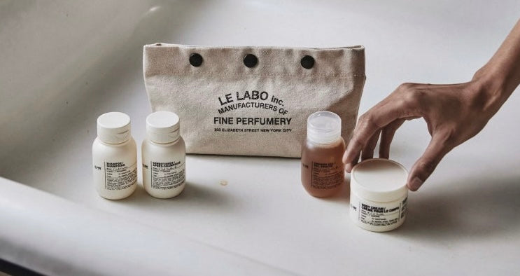 Le Labo self care product bottles on a white surface with a hand reaching for one of the bottles.