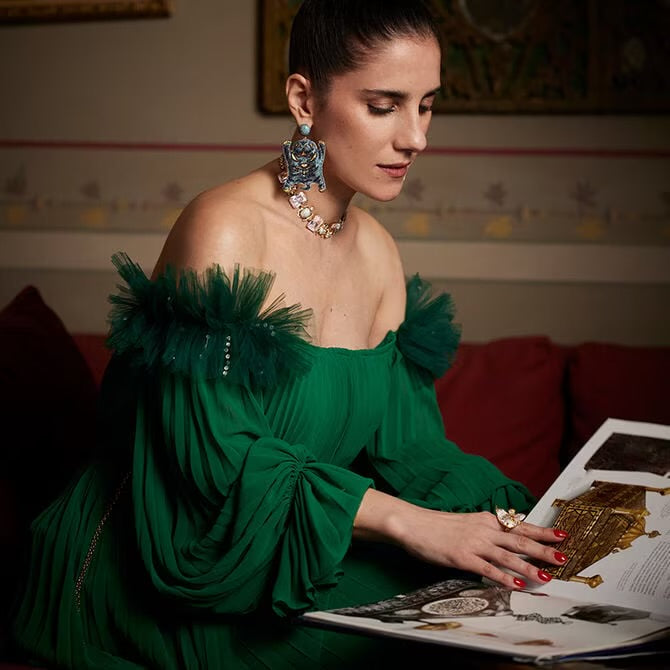  BEGÜM KHAN  in a green dress with feathered shoulders, sitting on a couch and looking at a magazine.