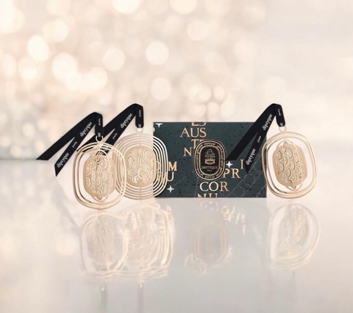 Diptyque decorative ornaments and a perfumed notebook, displayed with patterned paper packaging on a golden, sparkling background