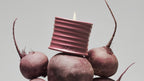 Loewe candle in a ribbed pink ceramic holder placed on beetroots, artistic composition with natural elements on a neutral background
