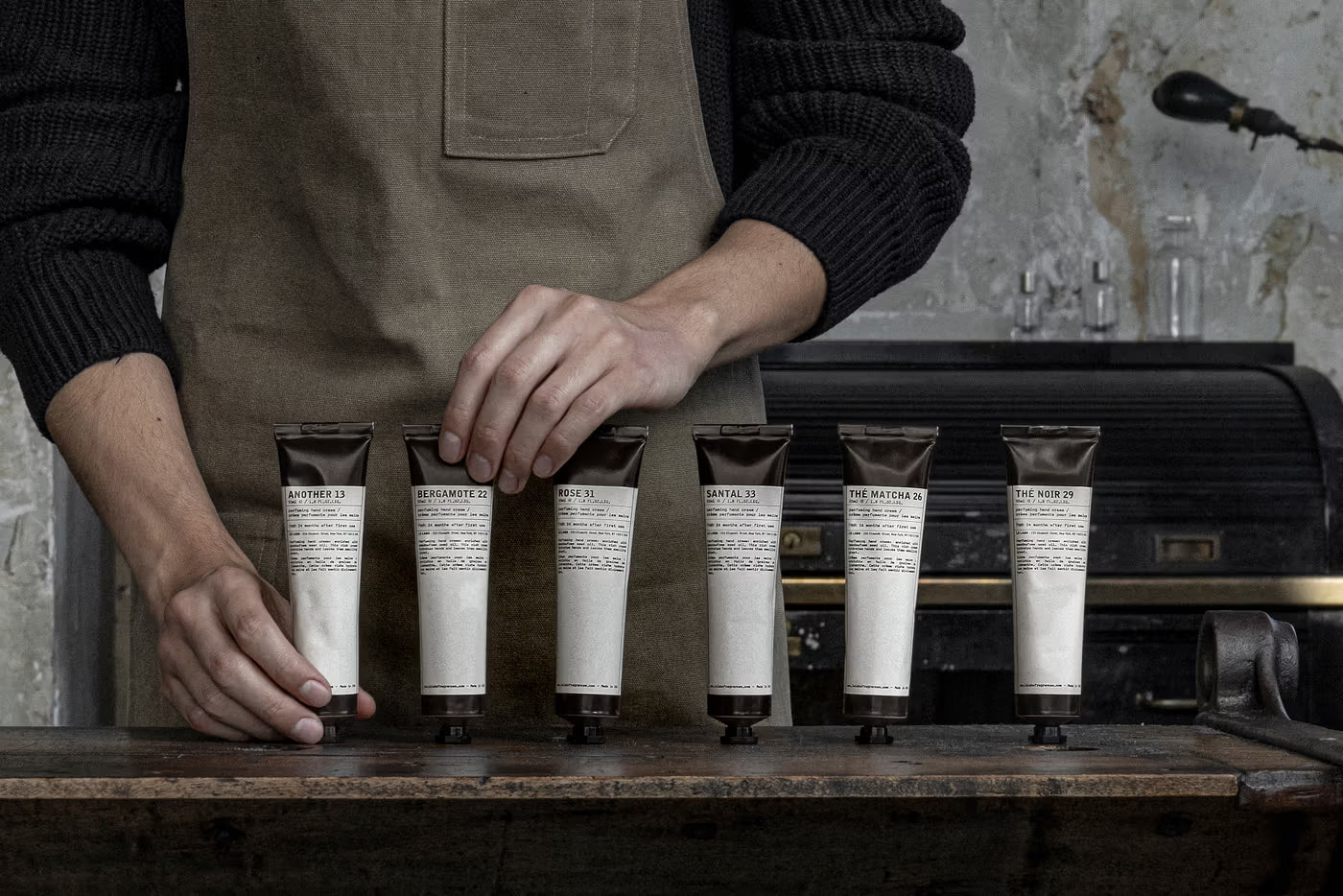 Person in a brown apron arranging small bottles on a wooden counter. le labo