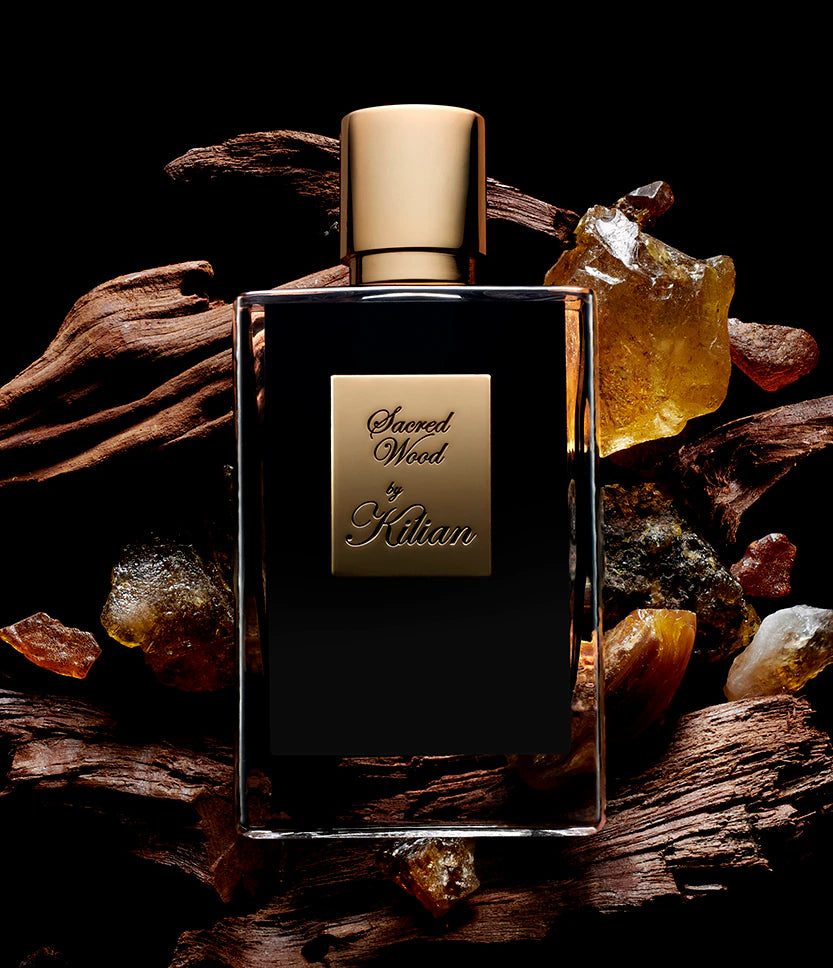 Perfume bottle labeled 'Sacred Wood by Kilian' on a dark background with amber stones and wood.